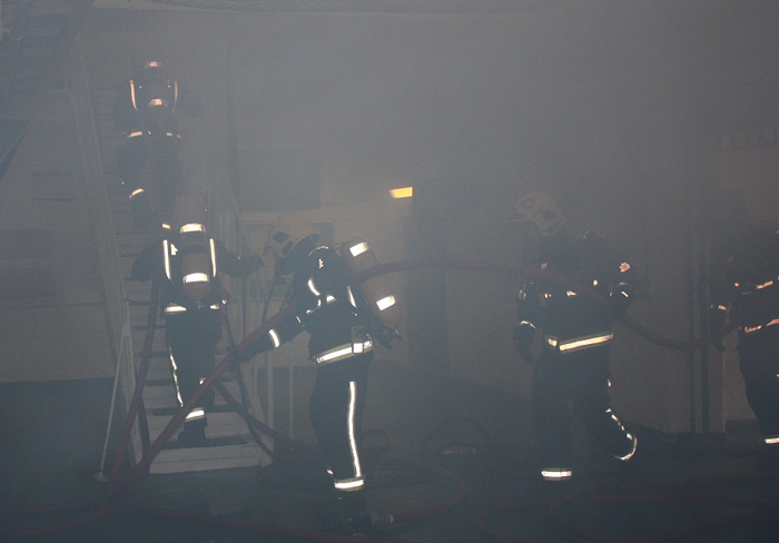 six firefighters can hardly be seen as they carry a hose through smoke and up a flight of stairs