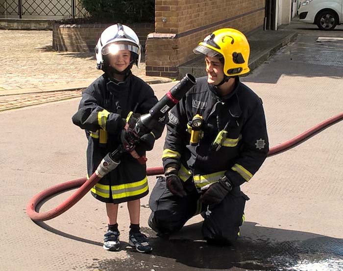 Young boy in a firefighters jacket and helmet stood next to a kneeling male firefighter 