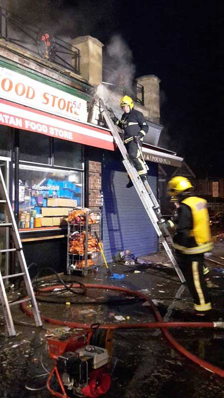 Two firefighters at a shop that is on fire, one is climbing a small ladder while the other looks at debris on the floor 