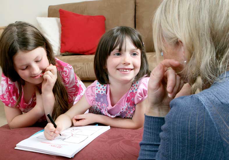 Two young girls drawing while  smiling at a family member