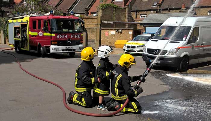 Young boy in a firefighters helmet and jacket stood with two firefighters as they use a hose