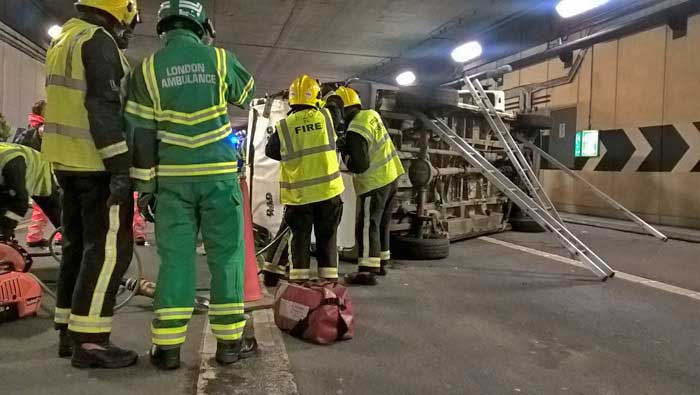 London Ambulance Service staff and firefighters in front of the overturned truck, ladders are propping the vehicle up