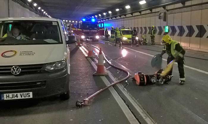 A van is facing straight ahead there is a long hose next to it with a fire engine with its lights on in the background 