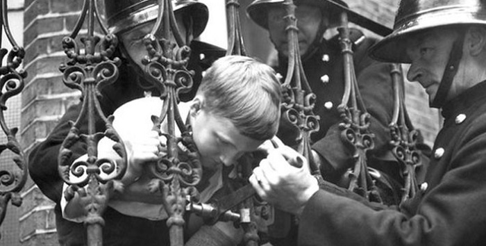 Black and white photo of firefighters rescuing a boy with his head stuck in the railings