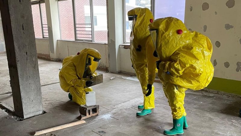 Three firefighters in yellow gas tight suits inspect a container of chemicals during a training exercise