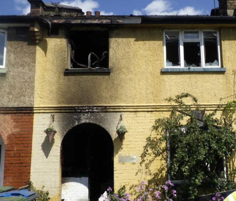 Front of house. Windows are broken and charred. Burnt debris sits outside the building.