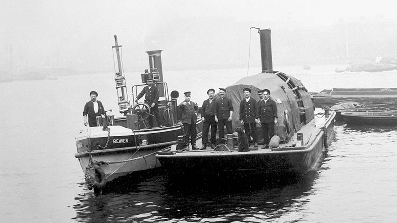 A group of Victorian firefighters stand on a firefighting barge, next to its tugboat 'Beaver'