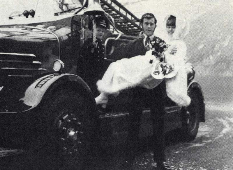 A man is lifting up a woman, they are a Bride and Groom. There is also a small boy. They are either standing on or near to a historical fire engine. Known as a ‘Big 4’ Braidwood-Bodied Fire Engine Pump, manufactured by Dennis around 1941.