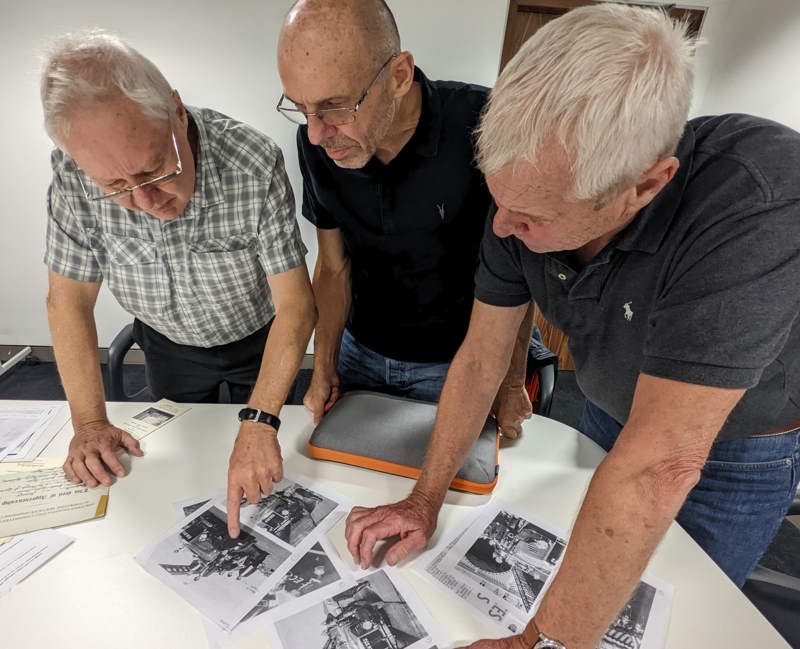 Three men are looking at printed copies of historical images, on a table. One man is point to a specific image.