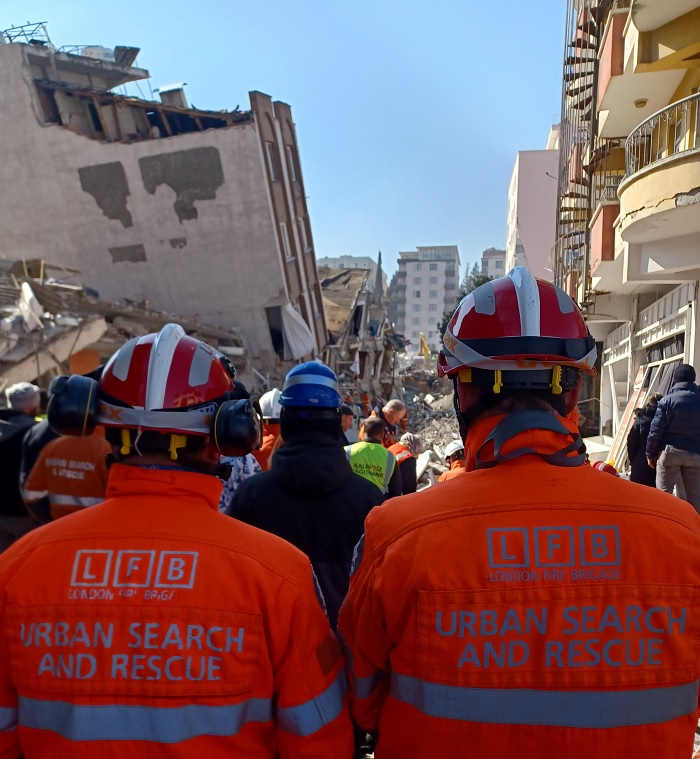 Two firefighters with their backs to the camera wearing hi-vis jackets with LFB Urban Search and Rescue on the back. Devastation from an earthquake is in the background.