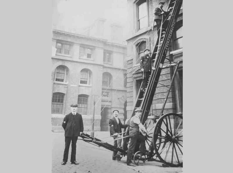 Firefighters from the Peek Frean Fire Brigade carrying out a fire drill in 1905. Two men are up the extended ladder, with a length of fire hose draped over the right shoulder of one of them. Three men stand at the bottom of the ladder, two are leaning on the ladder and the other stands nearby.