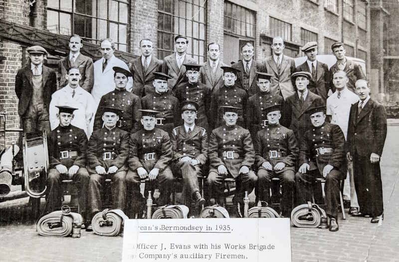 Group image of auxiliary firefighters from the Peek Frean Fire Brigade in 1935, at Bermondsey. Led by Officer J Evans. Some men are wearing the firefighter uniforms and have rolls of fire hose placed on the ground in front of them