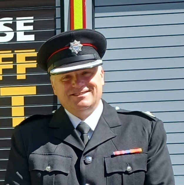 Borough Commander Richard stands in front of a fire engine. He is wearing his uniform and is illuminated by sunlight. 