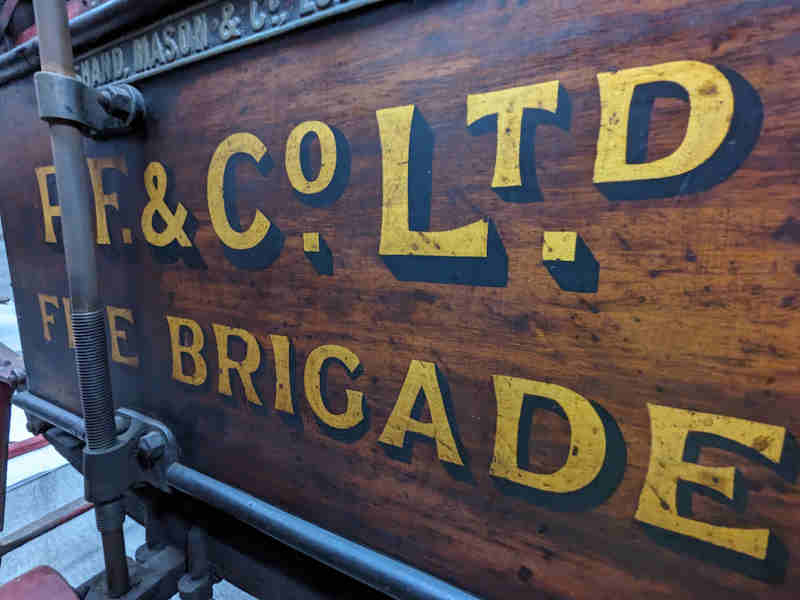 Close up of the central wooden storage section of the wheeled escape ladder from Peek Frean Fire Brigade. The Brigade's name is painted onto the side in yellow with black shadow style accents.